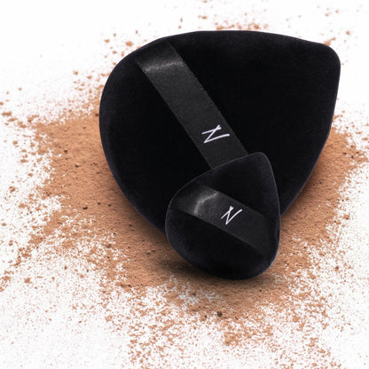 Close-up of the Velvet Powder Puff Duo, showing two luxurious black puffs with velvet texture on a backdrop of scattered powder.