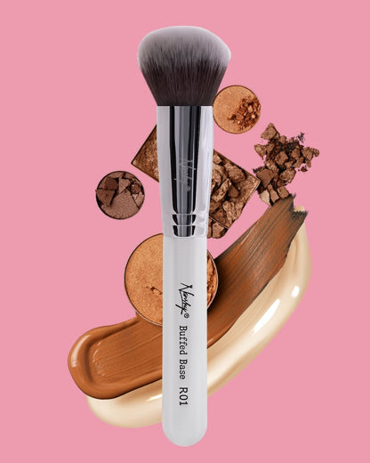 Nanshy Buffed Base makeup brush with soft bristles, surrounded by crushed and smeared makeup against a pink background.