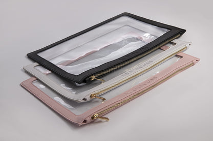 Stacked transparent PVC makeup pouches with 100% vegan leather edging in black, white, and pink, featuring golden brass zippers.